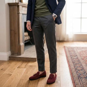 May include: A person wearing a navy blue blazer, olive green sweater, and gray trousers. The outfit is completed with burgundy loafers with tassels. The watch on the wrist has a silver band and face. The setting is a room with hardwood floors and a patterned rug.