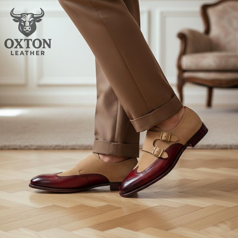 May include: Tan and burgundy leather double monk strap shoes with a wingtip design. The shoes feature a suede upper and polished leather toe and heel. Gold-coloured buckles secure the straps. The Oxton Leather logo is visible in the upper left corner.
