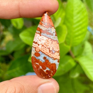 Può includere: Una gemma a forma di goccia con un motivo unico di vortici e macchie rosse, bianche e grigio-blu. La pietra lucida è tenuta tra le dita, mostrando i suoi colori e le sue trame naturali. Un esempio sorprendente di arte in pietra naturale.