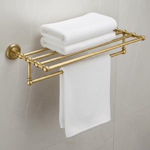 Puede incluir: Un toallero dorado montado en la pared con un diseño decorativo. El estante contiene dos toallas blancas dobladas en la parte superior y una toalla blanca colgando en la parte inferior. El estante está montado en una pared de azulejos gris claro.