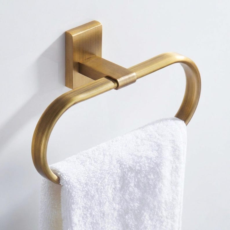 Peut inclure: Un porte-serviettes de couleur laiton fix&eacute; &agrave; un mur blanc, tenant une serviette blanche pli&eacute;e. L'anneau a une plaque de montage carr&eacute;e et une conception incurv&eacute;e et ouverte. La serviette est moelleuse et propre.