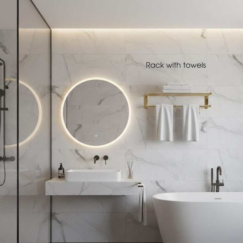 May include: A modern bathroom with a round mirror with a glowing edge, a white sink, and a gold towel rack with white towels. The text "Rack with towels" is visible. The walls are white marble.