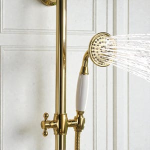 May include: Close-up of a gold shower fixture with a white handle. The showerhead is spraying water. The fixture is mounted on a white panelled wall.