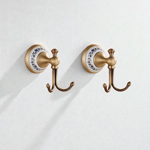 May include: Two antique-style wall hooks. Each hook has a circular, cream-colored base with a blue floral design. The hooks are made of bronze-colored metal and have a double hook design, perfect for hanging towels or robes.