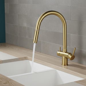 May include: A gold-colored kitchen faucet with a curved spout is shown with water flowing into a white double sink. The faucet is mounted on a light wood countertop, with a gray tile backsplash in the background.