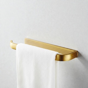 Könnte beinhalten: Ein goldfarbener Handtuchhalter mit einem weißen Handtuch. Der Halter ist an einer hellen Wand befestigt. Das Handtuch ist gefaltet und hängt herunter. Der Halter hat ein gebogenes Design.