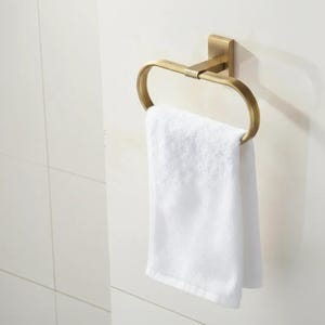 Peut inclure: Un porte-serviettes en m&eacute;tal dor&eacute; avec une serviette blanche suspendue. L'anneau est mont&eacute; sur une base carr&eacute;e, et la serviette est pli&eacute;e proprement. L'accessoire de salle de bain est contre un mur carrel&eacute; blanc.