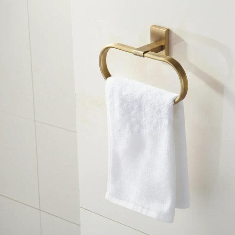 Peut inclure: Un porte-serviettes en m&eacute;tal dor&eacute; avec une serviette blanche suspendue. L'anneau est mont&eacute; sur une base carr&eacute;e, et la serviette est pli&eacute;e proprement. L'accessoire de salle de bain est contre un mur carrel&eacute; blanc.