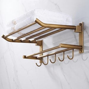 Op de afbeelding: Een aan de muur gemonteerde handdoekenrek van antiek messing met een plank met opgevouwen witte handdoeken. Onder de plank bevinden zich twee horizontale staven en vijf haken om handdoeken op te hangen. Gemonteerd op een wit marmerpatroon muur.