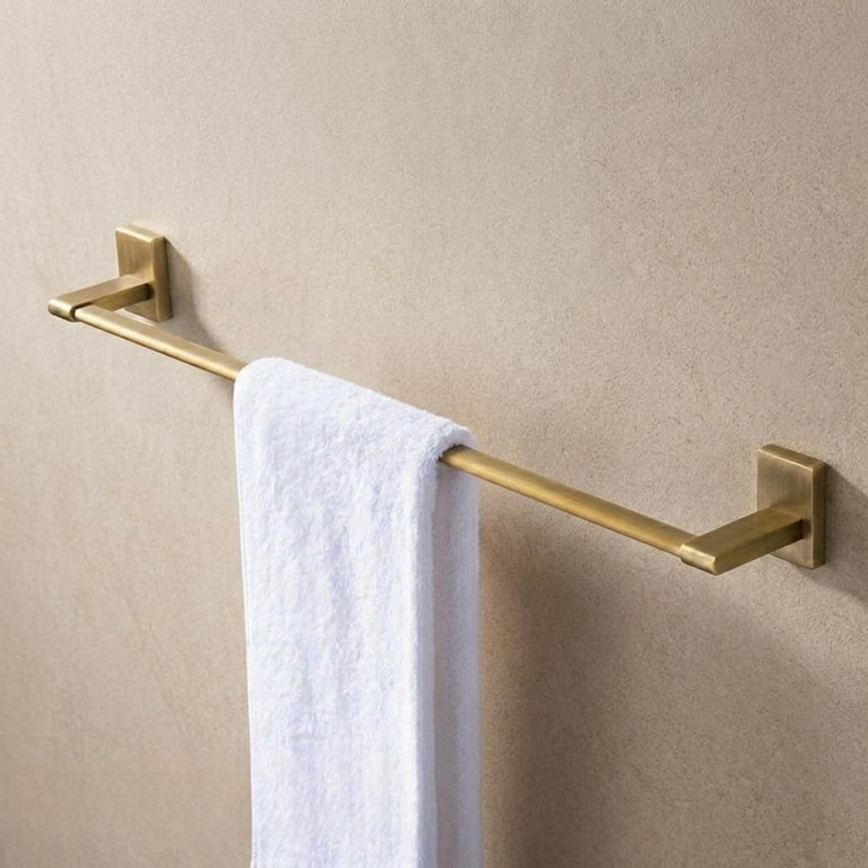 Peut inclure: Un porte-serviettes dor&eacute; fix&eacute; au mur, tenant une serviette blanche. La barre a un design rectangulaire avec des plaques de montage carr&eacute;es. La serviette est pli&eacute;e sur la barre. Le mur est de couleur marron clair neutre.