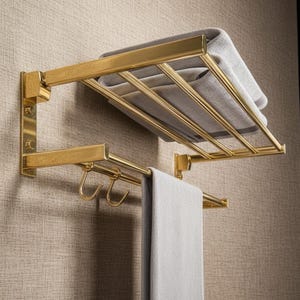 Puede incluir: Un toallero de metal dorado con un estante y una barra con ganchos. Una toalla gris doblada descansa sobre el estante, y otra toalla gris cuelga de la barra. El toallero está montado en una pared texturizada.