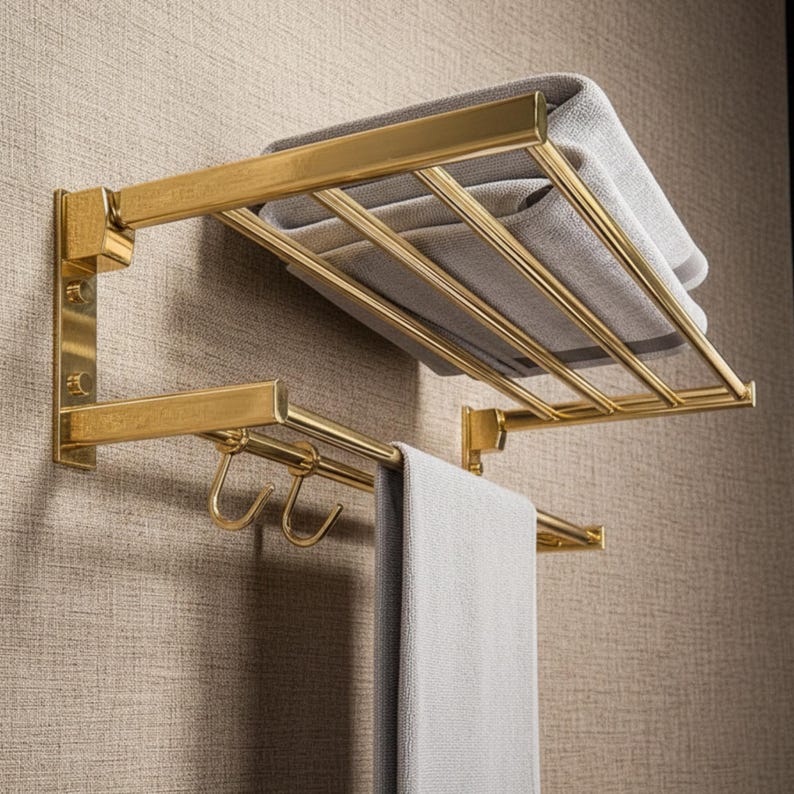 May include: A gold-colored metal towel rack with a shelf and a bar with hooks. A folded gray towel rests on the shelf, and another gray towel hangs from the bar. The rack is mounted on a textured wall.