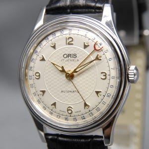 Puede incluir: Un reloj de pulsera automático Oris con esfera plateada, centro texturizado, manecillas doradas y números arábigos en 3, 6, 9 y 12. Una manecilla de fecha con punta de media luna roja indica la fecha en el borde exterior.