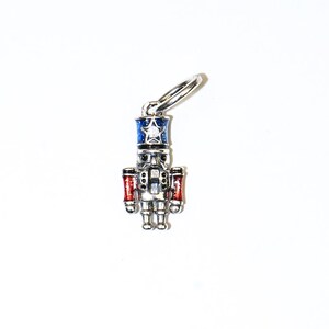 May include: A silver-colored charm in the shape of a nutcracker soldier. The charm has a blue hat with a white star, red arms, and a detailed face. The charm is attached to a silver ring.