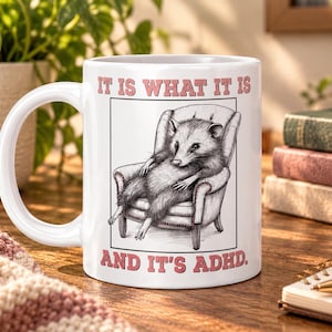 May include: A white ceramic mug featuring a black and white illustration of an opossum sitting in an armchair. The text above the illustration reads IT IS WHAT IT IS and the text below reads AND IT'S ADHD in pink capital letters.