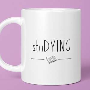 May include: White ceramic mug featuring the word "STUDYING" in black script, a line, and a small open book illustration. The mug has a curved handle and is set against a purple background.