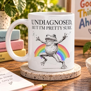 May include: A white ceramic mug featuring a black line drawing of a frog with arms outstretched in front of a colourful rainbow. The text above the frog reads, "UNDIAGNOSED, BUT I'M PRETTY SURE." The mug has a curved handle and sits on a speckled coaster.