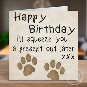 May include: A birthday card with the message "Happy Birthday I'll squeeze you a present out later" in black. Two brown paw prints are at the bottom. The card is on a wooden surface.