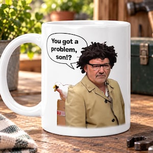 May include: A white ceramic mug featuring a graphic of a person with dark curly hair and glasses wearing a tan jacket. A white duck is visible behind a cardboard box. A speech bubble contains the black text: You got a problem, son!?