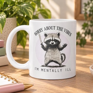 May include: A white ceramic mug featuring a black and white illustration of a raccoon holding a small sword. The text above the raccoon reads SORRY ABOUT THE VIBES and the text below reads I'M MENTALLY ILL. The mug has a curved handle.