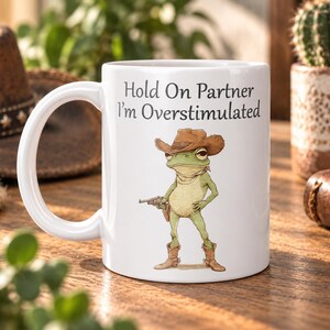 May include: A white ceramic mug featuring a graphic of a green frog wearing a brown cowboy hat and boots, holding a small revolver. The text above the frog reads, "Hold On Partner I'm Overstimulated" in a black, serif font centred on the mug.