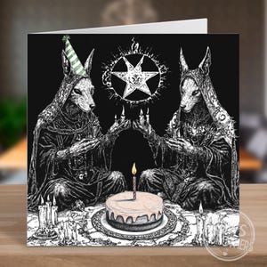 May include: Greeting card featuring a black and white illustration. Two figures with wolf-like heads in hooded robes sit either side of a cake with a lit candle. A pentagram and candles surround the cake. One figure wears a party hat.
