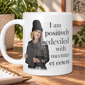 May include: A white ceramic mug featuring a graphic illustration of a person wearing a black hat and patterned top, holding a black folder. The text reads, I am positively bedeviled with meetings et cetera, printed in black serif font on the side.