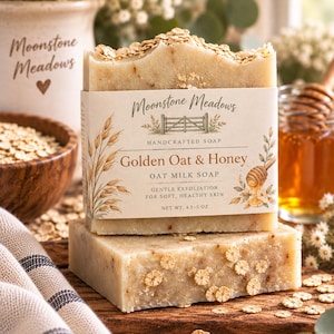 Könnte beinhalten: Ein rechteckiges Stück Hafermilchseife Golden Oat & Honey mit einem Papieretikett, auf dem Moonstone Meadows steht. Die Seife ist mit Haferflocken bestreut und an der Seite mit kleinen Trockenblumen verziert. Das Etikett gibt ein Nettogewicht von 4,5-5 oz an.