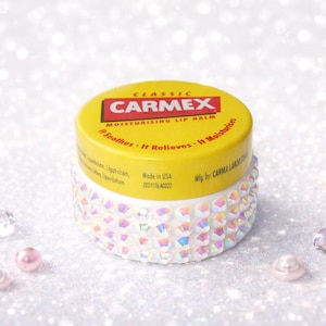 Könnte beinhalten: Ein gelb-weißes Carmex Lippenbalsam-Glas mit einer verzierten Basis. Der Deckel zeigt das Carmex-Logo und Produktinformationen. Der Hintergrund ist eine glitzernde Oberfläche mit verstreuten Schmucksteinen.