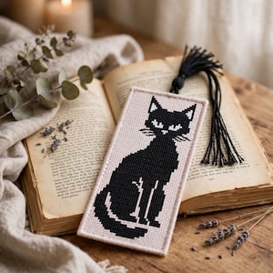 Peut inclure: Un marque-page rectangulaire orné d'un motif au point de croix représentant un chat noir sur un fond en tissu beige clair. Le marque-page est terminé par un pompon en fil noir fixé au bord supérieur, mettant en valeur une silhouette féline.