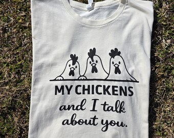 May include: Cream-colored t-shirt with a black graphic of three cartoon chickens and the text "MY CHICKENS and I talk about you." The shirt is laid flat on a bed of brown and green grass.