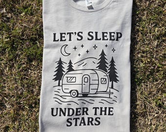 May include: A light gray t-shirt with the text "LET'S SLEEP UNDER THE STARS" in black. The design includes a camper, trees, a moon, and stars. The shirt is folded on a bed of grass.