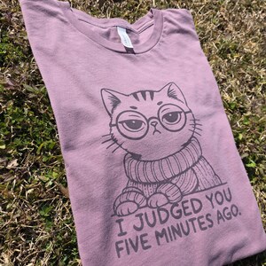May include: Mauve-colored t-shirt featuring a cartoon cat wearing glasses and a sweater. The cat has a judgmental expression. The text "I Judged You Five Minutes Ago" is printed below the cat.