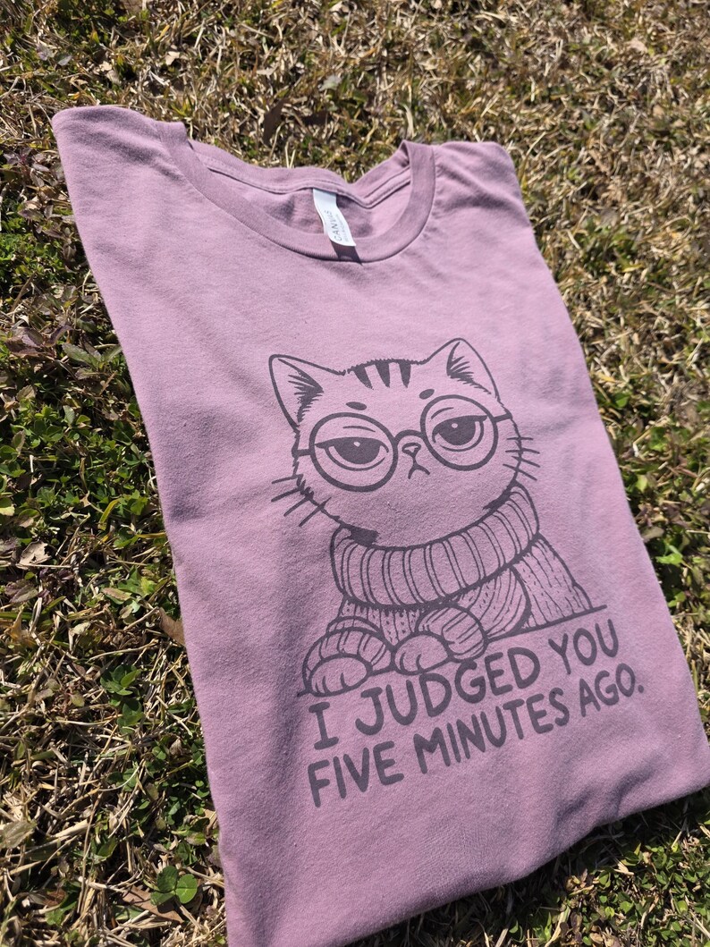 May include: Mauve-colored t-shirt featuring a cartoon cat wearing glasses and a sweater. The cat has a judgmental expression. The text "I Judged You Five Minutes Ago" is printed below the cat.