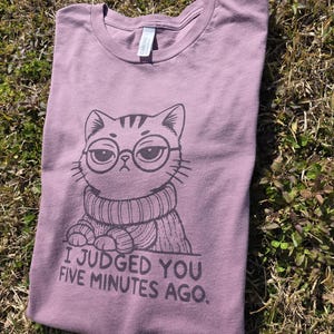 May include: Mauve t-shirt with a graphic of a cat wearing glasses and a sweater. The text "I JUDGED YOU FIVE MINUTES AGO" is printed below the cat illustration. The shirt is folded on a grassy surface.