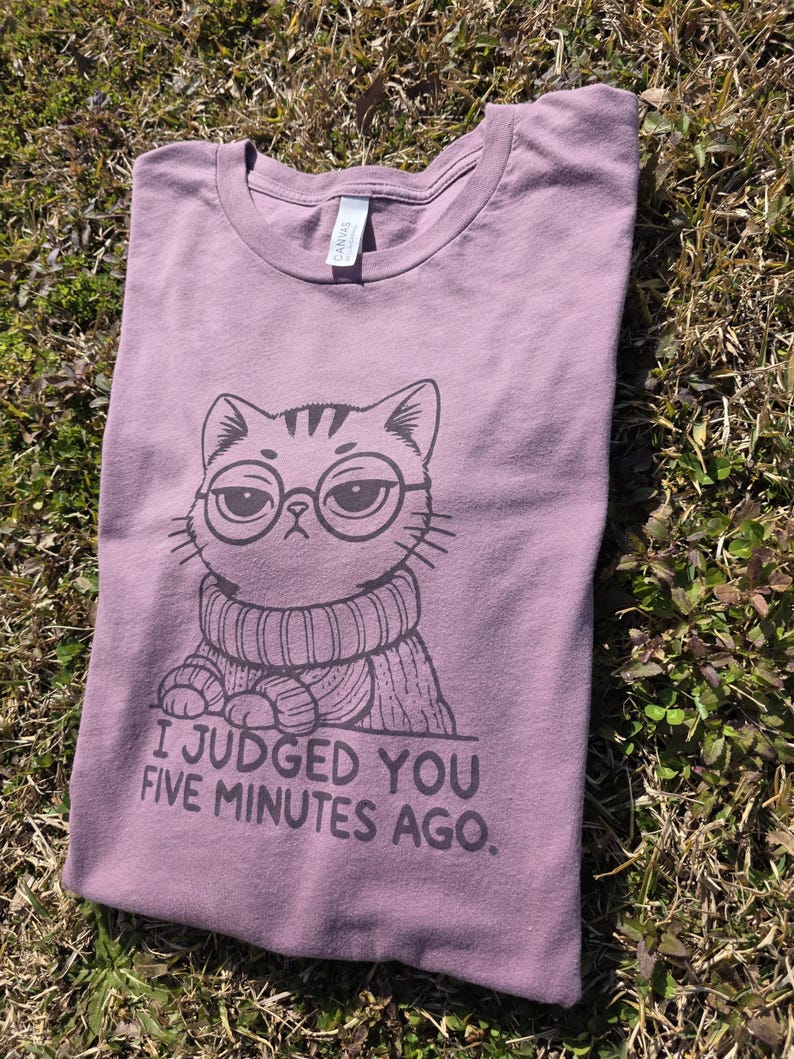 May include: Mauve t-shirt with a graphic of a cat wearing glasses and a sweater. The text "I JUDGED YOU FIVE MINUTES AGO" is printed below the cat illustration. The shirt is folded on a grassy surface.