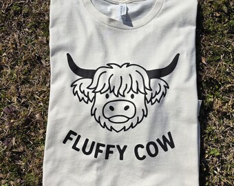 May include: A cream-colored t-shirt featuring a black graphic of a shaggy cow head with horns and the text "FLUFFY COW" printed in a bold, sans-serif font below the illustration. The shirt has a crew neckline and short sleeves.