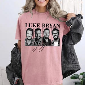 May include: Pink t-shirt featuring the text "LUKE BRYAN" above four black and white images of the singer. The shirt also includes a signature graphic in black.