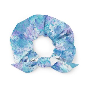Puede incluir: Una scrunchie con un diseño de acuarela en tonos azules, turquesas y morados. La tela está fruncida y atada en forma de lazo. Este accesorio para el cabello es una adición elegante a cualquier peinado.