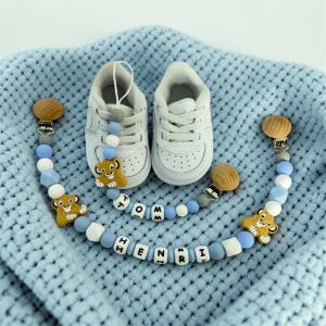 Könnte beinhalten: Zwei weiße Babyschuhe neben zwei Schnullerhaltern. Die Halterungen haben Holz- und Silberakzente, blaue und weiße Perlen sowie Löwen-Charms. Eine Halterung schreibt "TOMM" und die andere "HENRI". Die Artikel liegen auf einer blauen Decke.