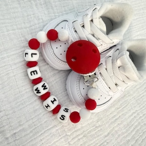 May include: A red and white pacifier clip with the name "LEWISH" spelled out in black letters on white beads. The clip is next to a pair of white baby trainers.