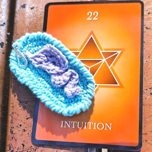 Op de afbeelding: Een handgemaakte, turquoise gehaakte teenslipper sleutelhanger met een lavendelkleurige band en zilveren sluiting ligt op een oranje kaart. De kaart toont het nummer 22, een geometrisch ontwerp en het woord "INTUITION" in het wit.