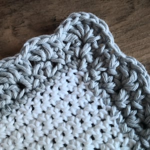May include: Close-up of a handmade crochet item with a scalloped edge. The item features a two-tone design, with a white center and a gray border. The stitches are clearly visible, showcasing the texture of the yarn.
