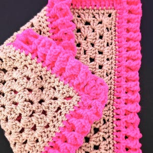 May include: A handmade crochet blanket with a tan central section and a bright pink border. The blanket has a textured stitch pattern and a ruffled edge, creating a cosy and decorative item. The blanket is folded, showing the contrasting colours and textures.