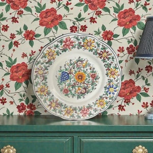 Könnte beinhalten: Ein dekorativer Keramikteller mit gewelltem Rand, der ein Blumenmuster in Gelb, Rosa, Blau und Orange aufweist. Der Teller steht vor einem floralen Tapetenhintergrund und erzeugt eine Vintage-Ästhetik.