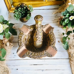 Peut inclure: Un cendrier vintage au design unique. Le cendrier est principalement doré avec une surface texturée et comporte quatre zones de repos. Le centre a une forme cylindrique. Le cendrier est posé sur une surface en bois blanc.