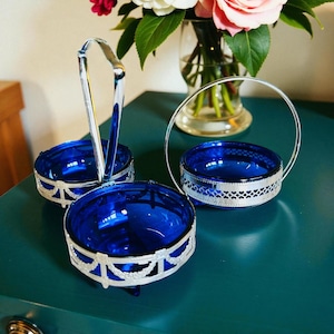 Peut inclure: Ensemble de trois coupelles en verre bleu cobalt avec des bordures et des poignées en métal argenté ornementé. Les coupelles sont disposées sur une surface turquoise, avec un vase de fleurs en arrière-plan. L'ensemble est probablement destiné au service ou à l'exposition.