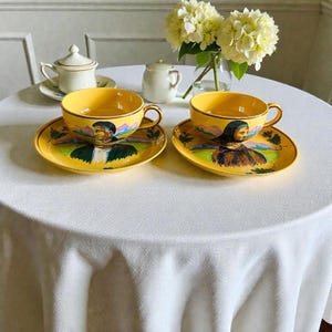 Peut inclure: Deux tasses et soucoupes jaunes avec des portraits peints, posées sur une nappe blanche. Les tasses et les soucoupes ont une couleur de base jaune avec un portrait peint d'une personne. Un sucrier et une crémière sont également sur la table.