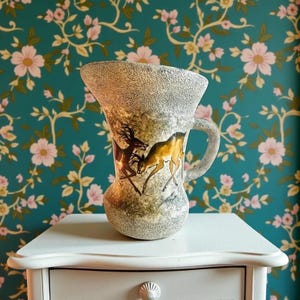May include: A vintage vase with a handle, decorated with a deer illustration in brown and gold. The vase has a textured surface and a wide opening. The background is a floral wallpaper with pink and white flowers.