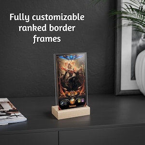 May include: A clear acrylic display frame with a colorful fantasy art print, set on a light wood base. The frame features the text "Fully customizable ranked border frames". The artwork has a woman in a black dress with red and gold accents.